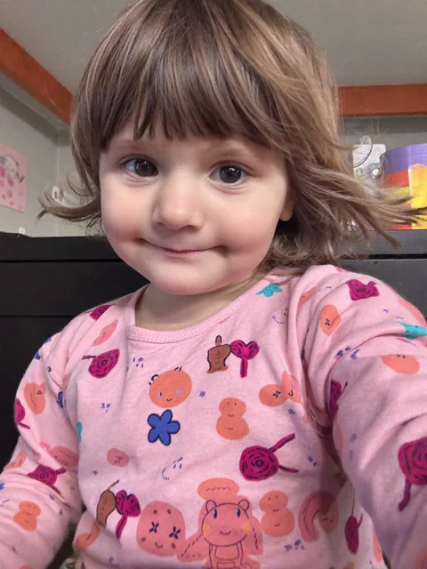 A smiling child with a brown bob and bangs, in a pink top covered in little drawings